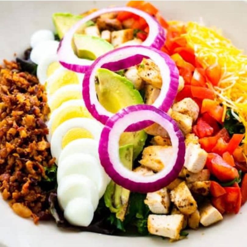 A vibrant Pujo St Cafe salad with chicken, avocado, red onion, egg, bacon, and cheese.